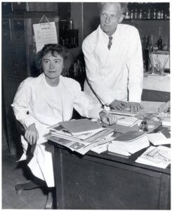 A woman sitting at a desk with a lot of papers and a man standing beside her